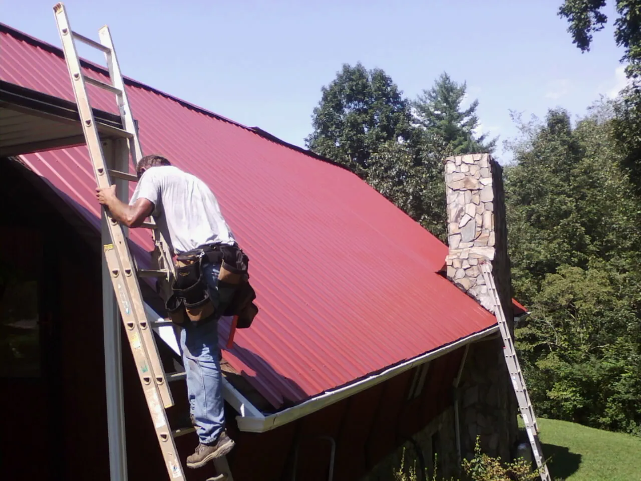 Roofer on a ladder beside a red metal roof and stone chimney — Soffit & Fascia Repair by Roofing Brothers in Renton